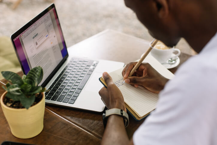 Rearview shot of interracial man using laptop, writing notes in textbook and drinking coffee in internet cafe with wireless technology. Student preparing to exam, use PC technology, remote education