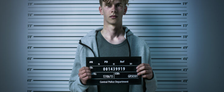 In a Police Station Arrested Drug Addict Teenage Posing for a Front View Mugshot. He is Heavily Bruised. Height Chart in the Background.